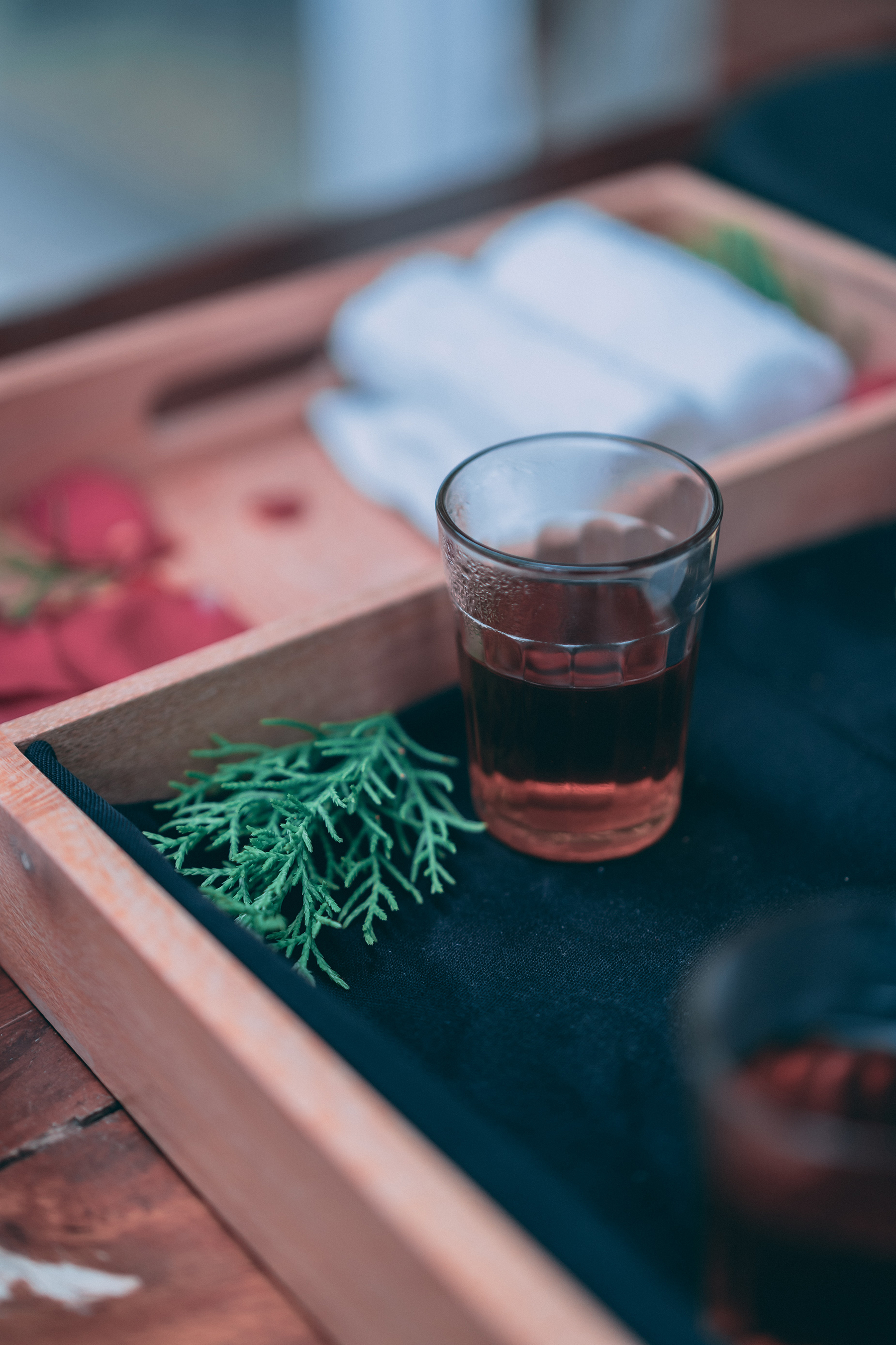 A glass of dark herbal tea on a wooden tray, with green leaves and towels in the background.